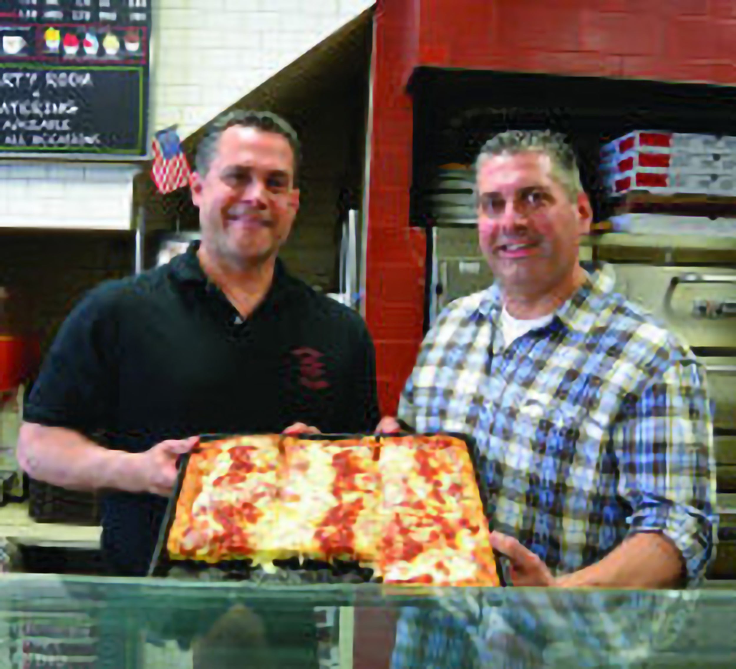 Two men holding a large square pizza in a kitchen setting.