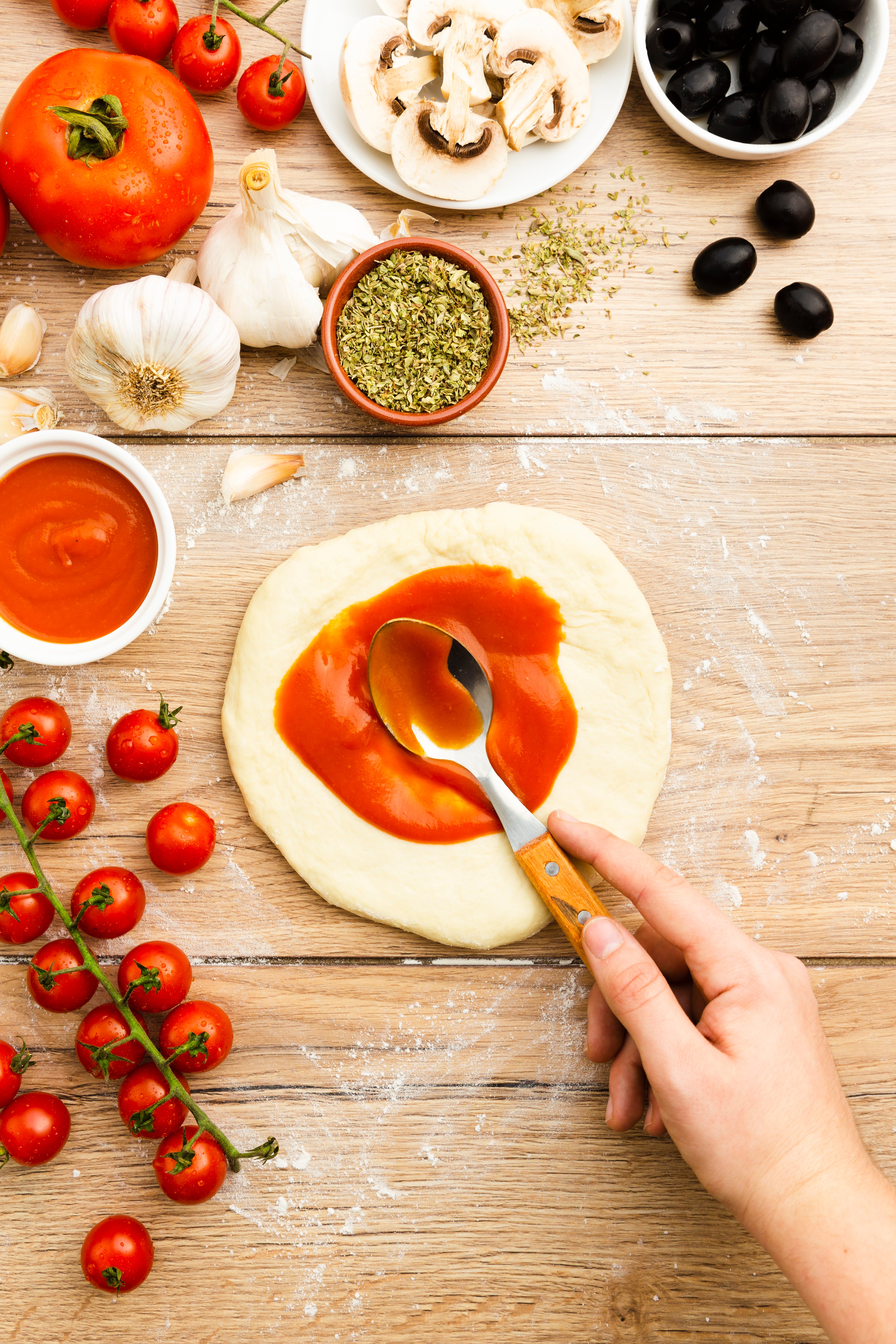 Person spreading tomato sauce on pizza dough with various ingredients around on a wooden surface